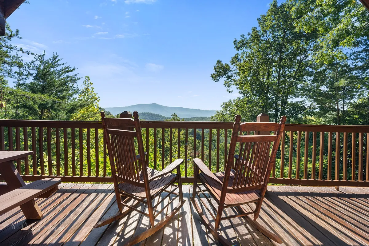 A wide wooden deck lined with classic rocking chairs faces out toward a lush, green mountain landscape. The foreground features comfortable seating perfect for relaxing, reading, or conversing, while the sweeping mountain views and greenery reinforce the peaceful atmosphere. Early morning or sunset light bathes the entire scene, evoking tranquility.