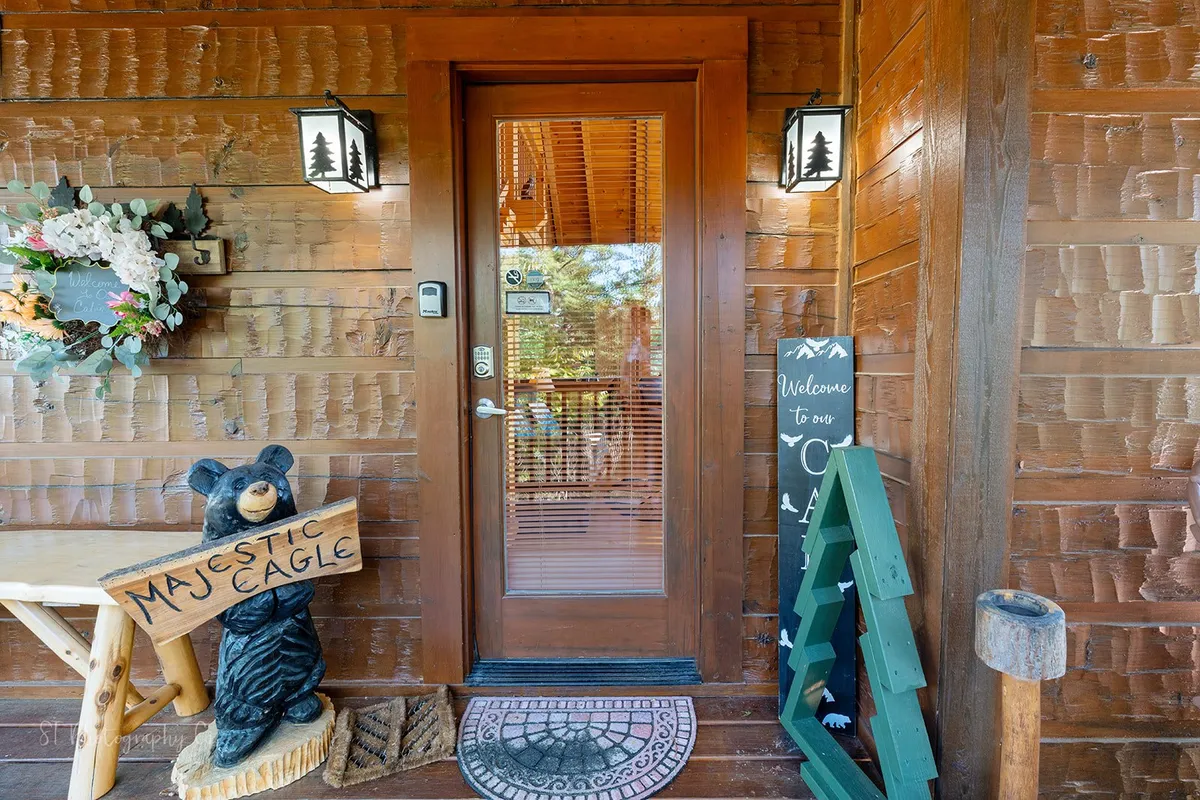 A welcoming entrance scene featuring a wooden door, bear statue, and a 'welcome' sign with a decorative wreath. The entrance is framed by rustic wood and surrounded by greenery, creating a sense of arrival and anticipation.