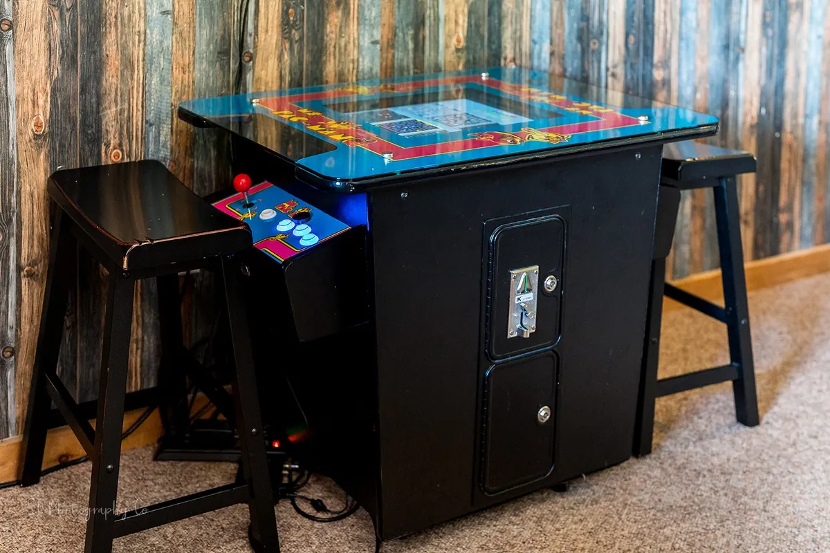 An inviting corner featuring a classic arcade game table with comfortable stools and warm wooden paneling, evoking a sense of nostalgia and fun. It's a perfect spot for engaging in friendly competitions and reliving arcade memories with friends or family.