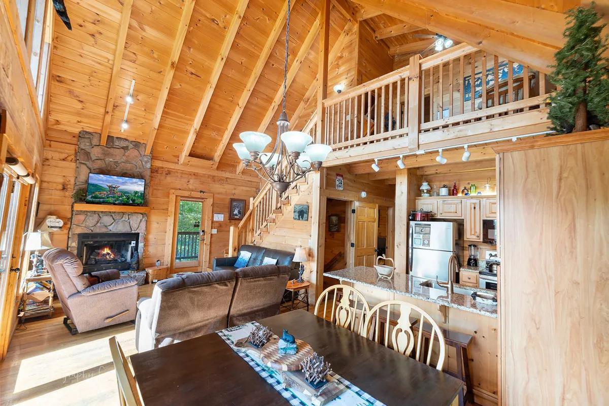 A spacious great room with a stone fireplace, an expansive dining table, a lofted balcony above, and large windows letting in plenty of natural light. The layout highlights both the dining and living areas, underscoring the open-plan design.
