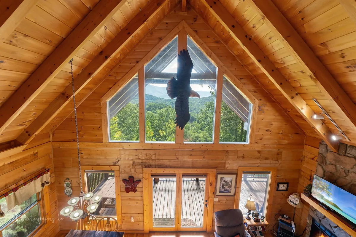 A large, light-filled common area with floor-to-ceiling windows framing breathtaking mountain views. A dining table and comfortable seating area are warmly lit by a chandelier, with rustic beams and a hanging eagle decor adding charm. The space radiates comfort and togetherness, ideal for team gatherings or brainstorming sessions.