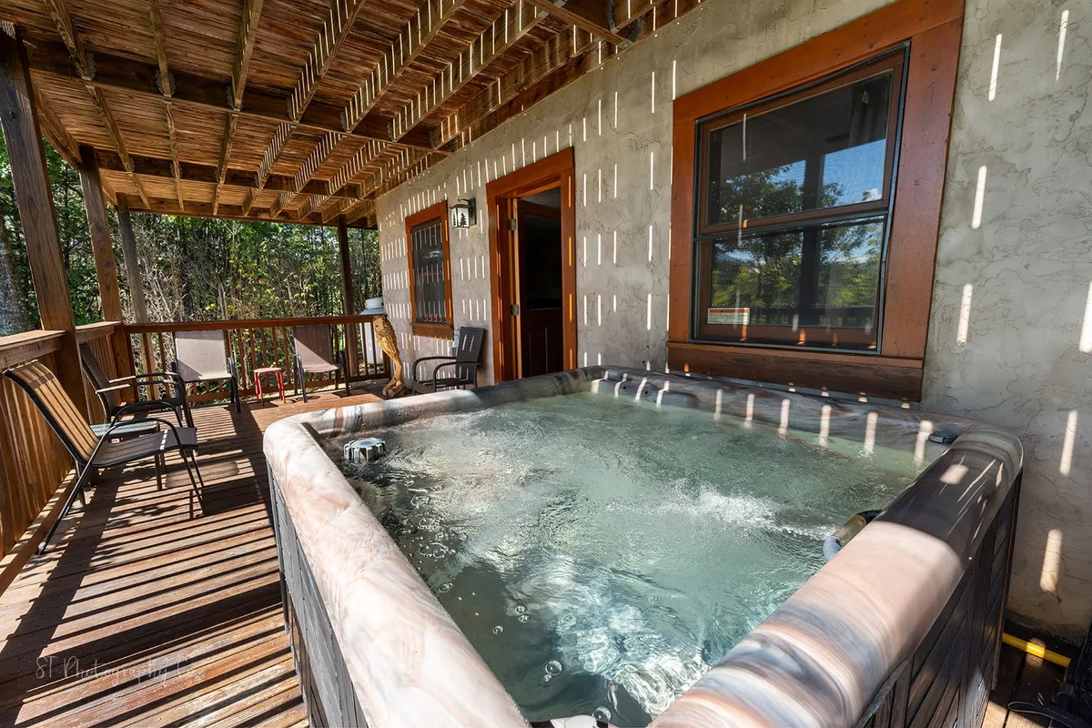 A bubbling hot tub is nestled on a wooden deck surrounded by lush greenery, offering a secluded and tranquil atmosphere. The setting is ideal for soaking and relaxing in nature, with the deck positioned for sunset views or stargazing.