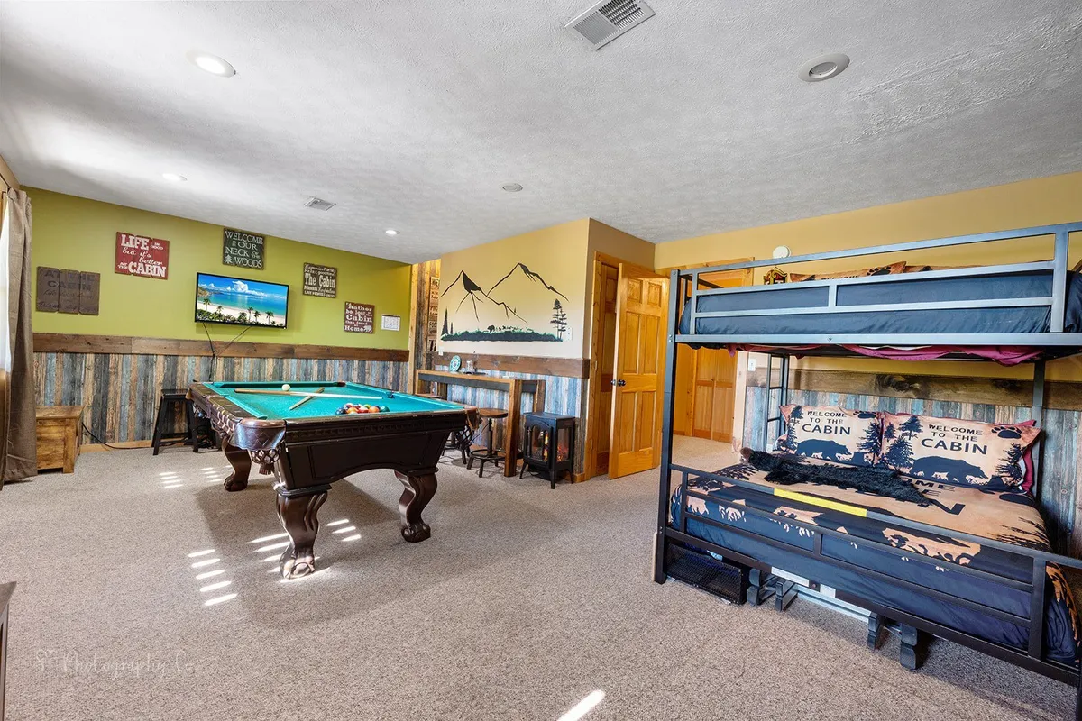 This image captures a vibrant game room featuring a pool table, queen-over-queen bunk beds, a large TV, mountain mural, and wooden wall accents. The setting exudes a fun, relaxed atmosphere where guests can play games, watch movies, socialize, or unwind on flexible sleeping arrangements.