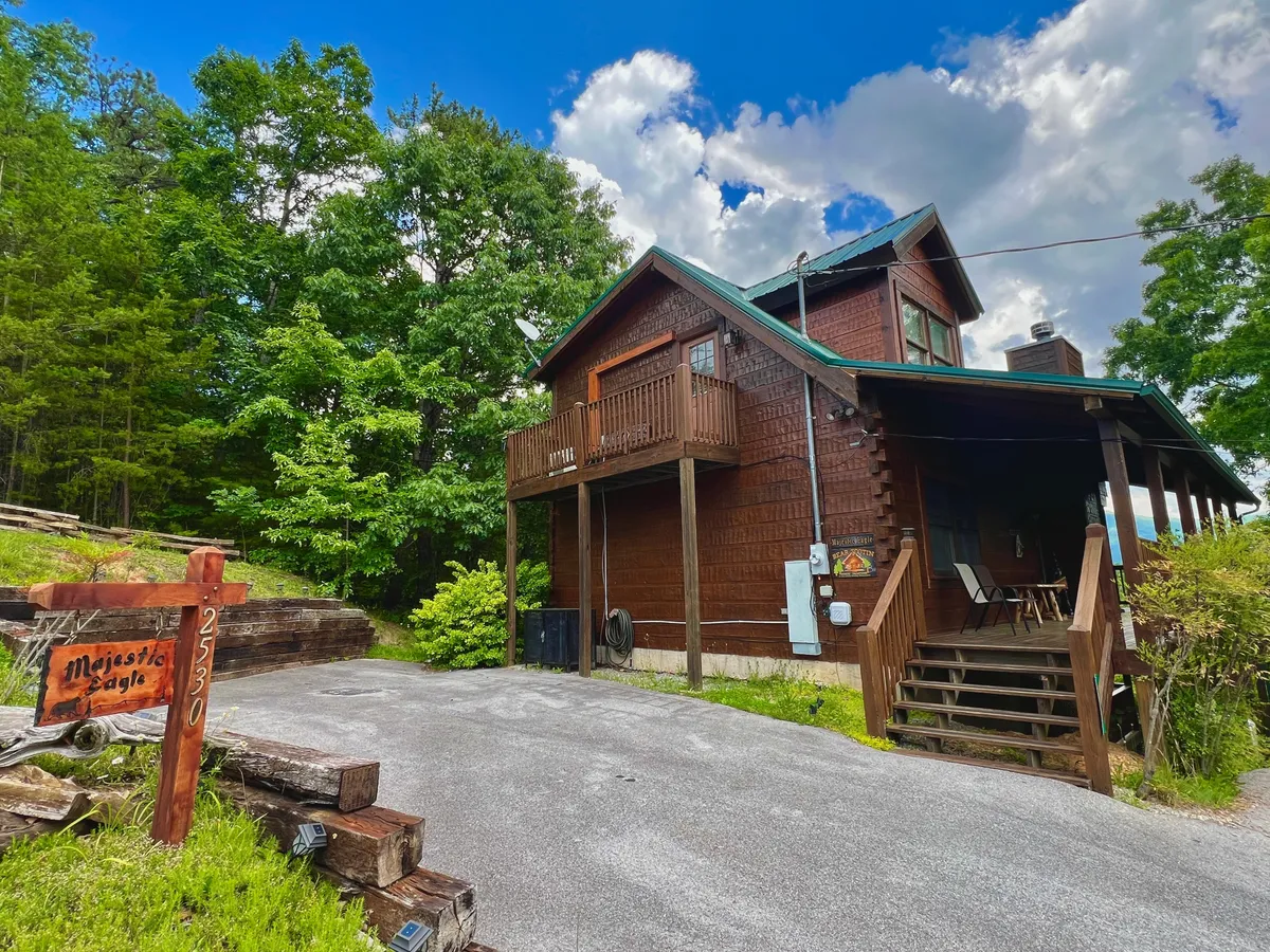 An exterior view of a wooden balcony with a signpost displaying the property's name. The balcony overlooks lush greenery and a stone pathway, suggesting a connection to the outdoors and providing a sense of place ideal for launching excursions.