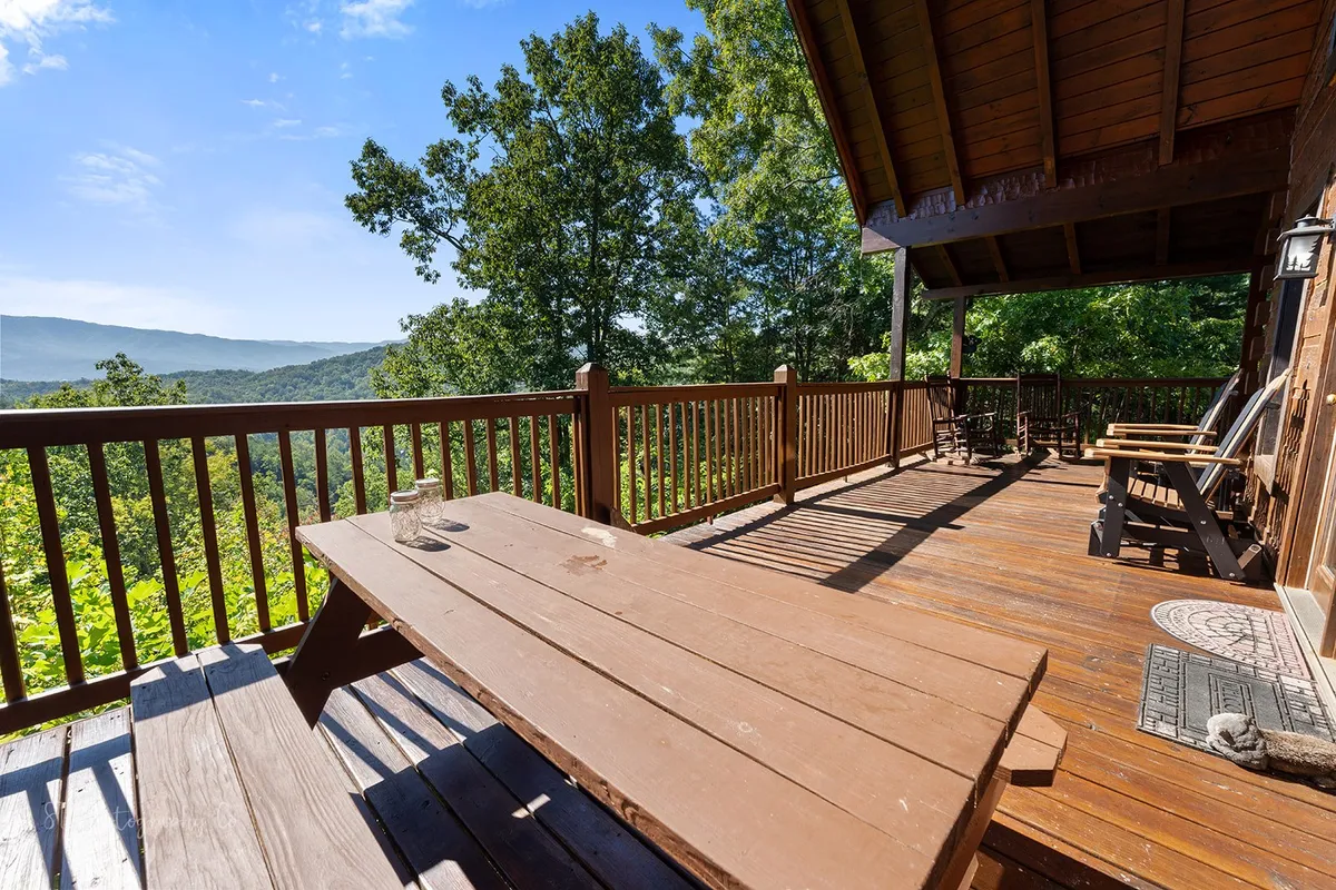 A rustic wooden deck with a picnic table and rocking chairs overlooks majestic mountain peaks and lush valleys. The deck is surrounded by sturdy wooden railings, inviting guests to dine, relax, or simply gaze at the breathtaking sunrise or sunset across the mountains.