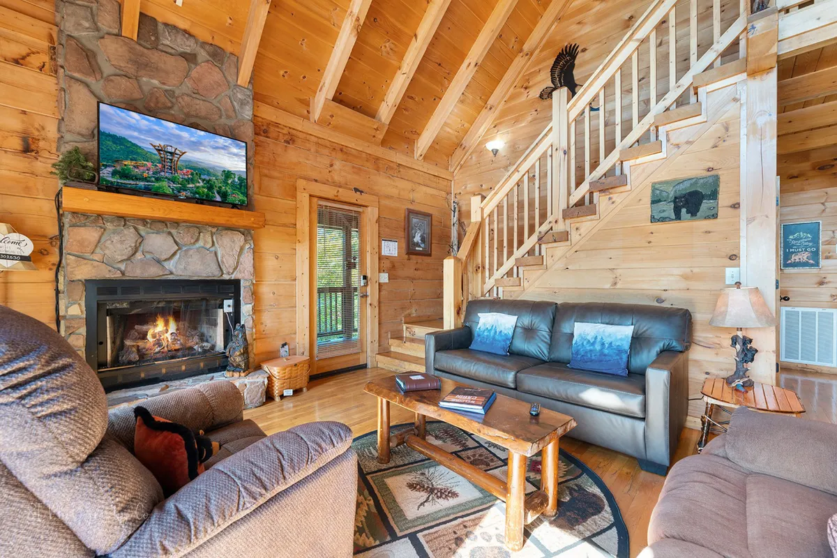 This image captures a cozy living space featuring a stone fireplace, multiple plush sofas, a television, and a staircase leading to an upper level. Light streams in from the windows, highlighting the inviting atmosphere and central gathering area.