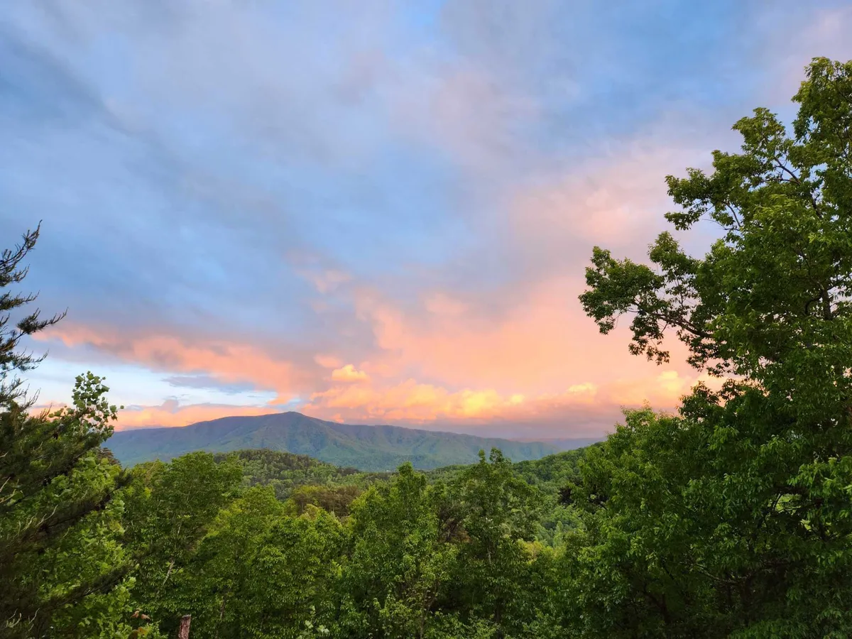 A stunning view of rolling mountains beneath a colorful sky, framed by lush greenery. The perspective suggests being immersed in the natural beauty of the region, highlighting the area's appeal for hikers, photographers, and outdoor enthusiasts across all seasons.