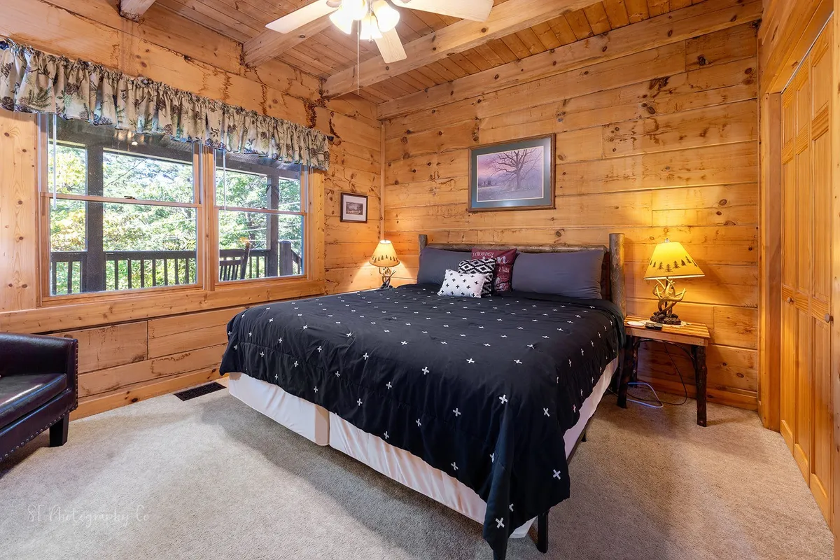 A tranquil bedroom showcasing a bed with black and white bedding, wooden walls, large windows offering beautiful views, and nightstands with lamps. The ambiance is peaceful and inviting, perfect for private relaxation.