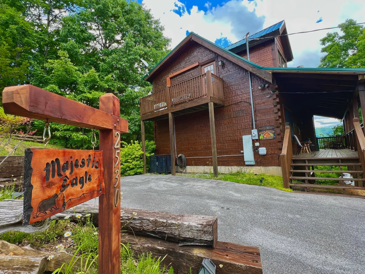 This exterior image features a wooden sign with 'Majestic Eagle' prominently displayed, the rustic wooden exterior of the house, and a balcony that overlooks a lush, green yard. The setting evokes a sense of arrival at a mountain retreat and hints at the natural adventures available just beyond the property.