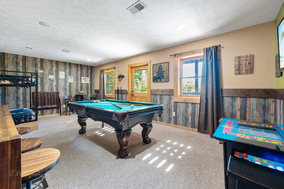 A bright, casual game space with a pool table, arcade game, wooden decor, large windows letting in natural light, and bunk beds. The room is arranged for friendly competitions, relaxation, and socializing, capturing both daytime and evening activities.